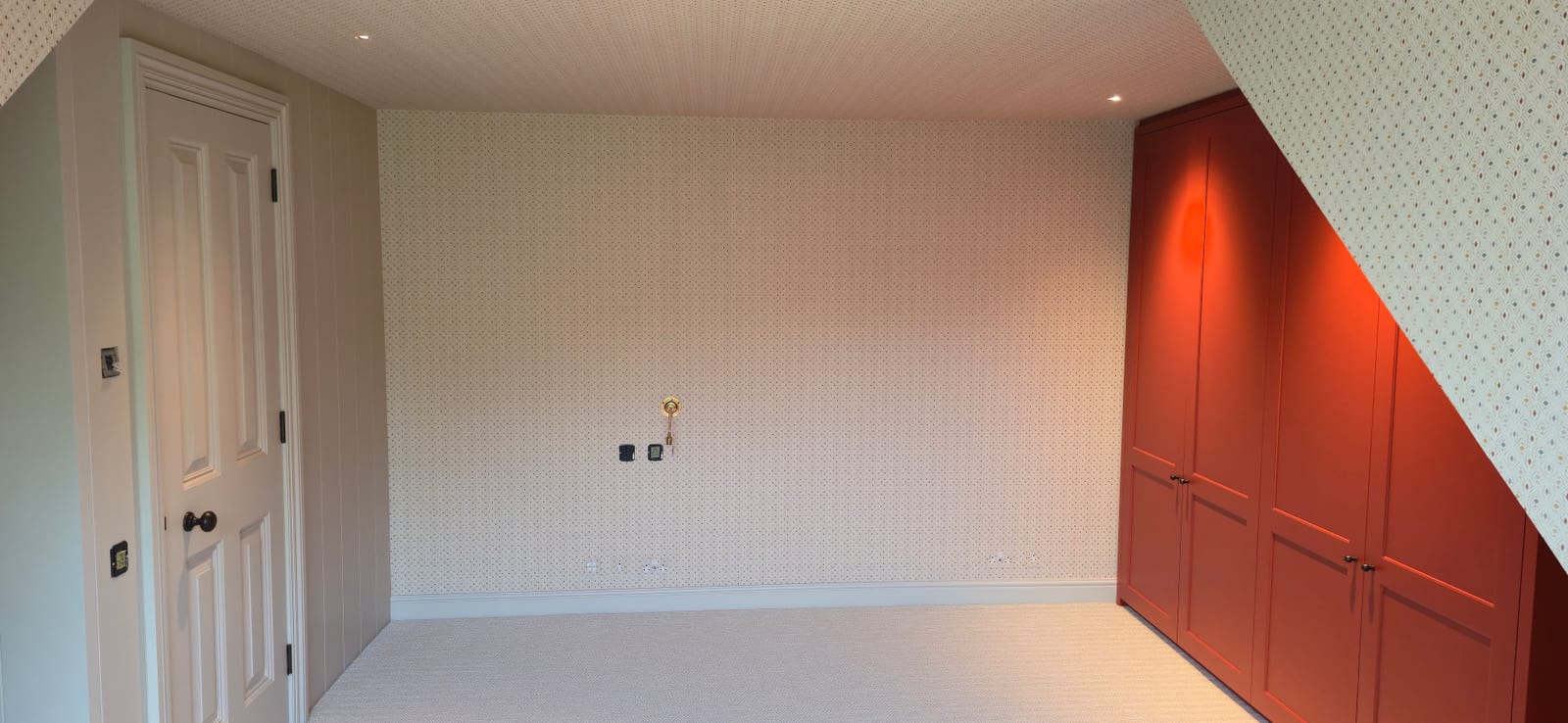 Attic bedroom with cream dotted wallpaper and terracotta built-in wardrobes under the eaves.